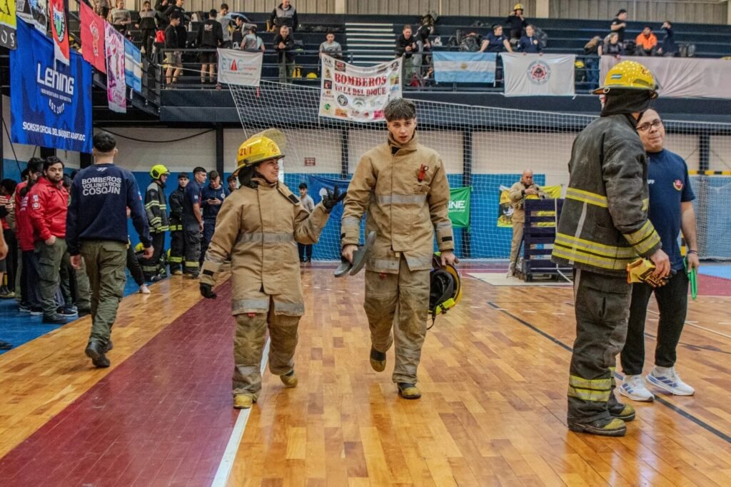 Bomberos equipados y público observan la jornada dentro de un gimnasio
