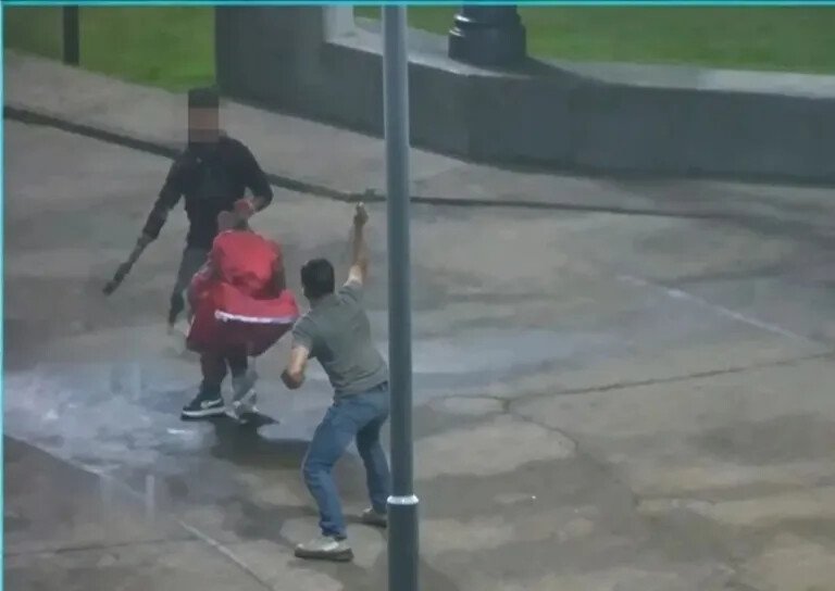Personas enfrentadas en una plaza; una sostiene un objeto alargado y otra lleva una prenda o mochila roja
