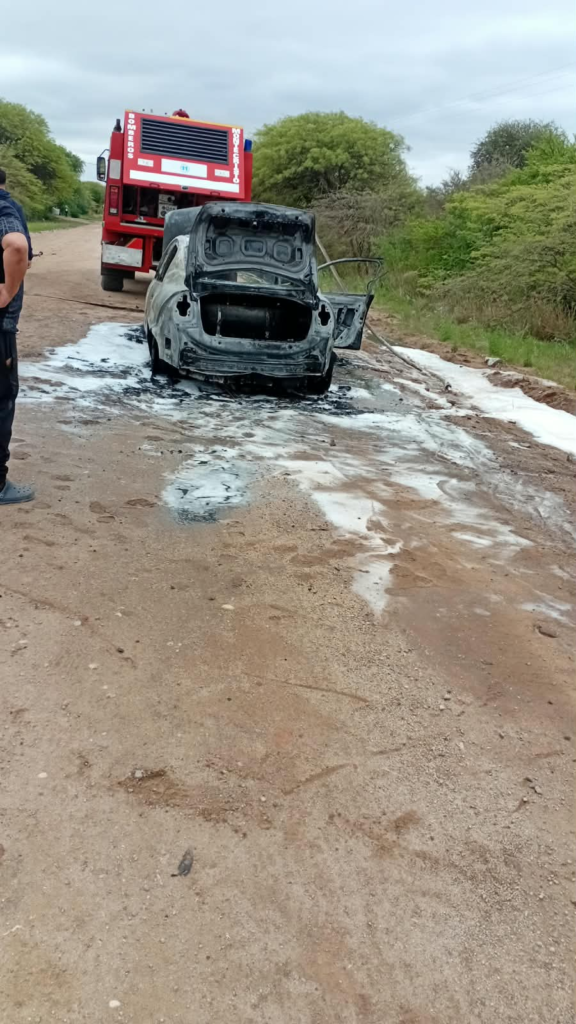 Automóvil incendiado sobre una calle de tierra, con una unidad de Bomberos Voluntarios detrás.